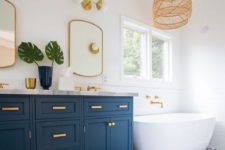 a coastal bathroom with blue and white printed tiles, a blue and gold vanity, gold frame mirrors, a woven lamp and a free-standing tub