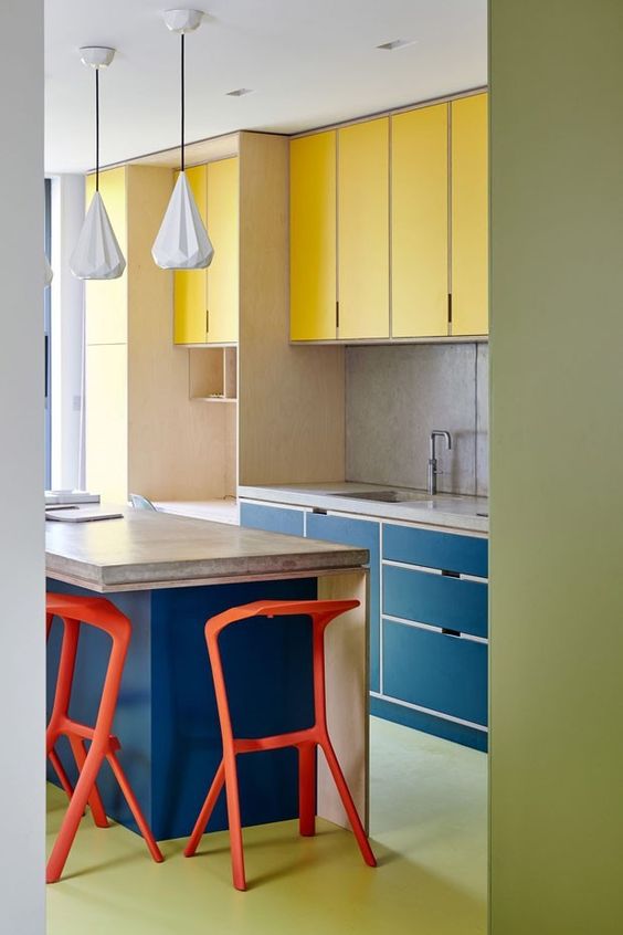 a colorful modern kitchen done in yellow and navy, with a stone countertop, red stools and a green floor is extra bold