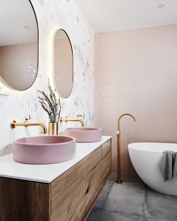a contemporary bathroom with blush hex tiles on the wall, pink sinks, glam fixtures and lit up gold frame mirrors