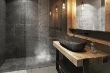 a creative grey bathroom done with grey tiles, a wood and metal vanity, a stone sink and a large lit up mirror