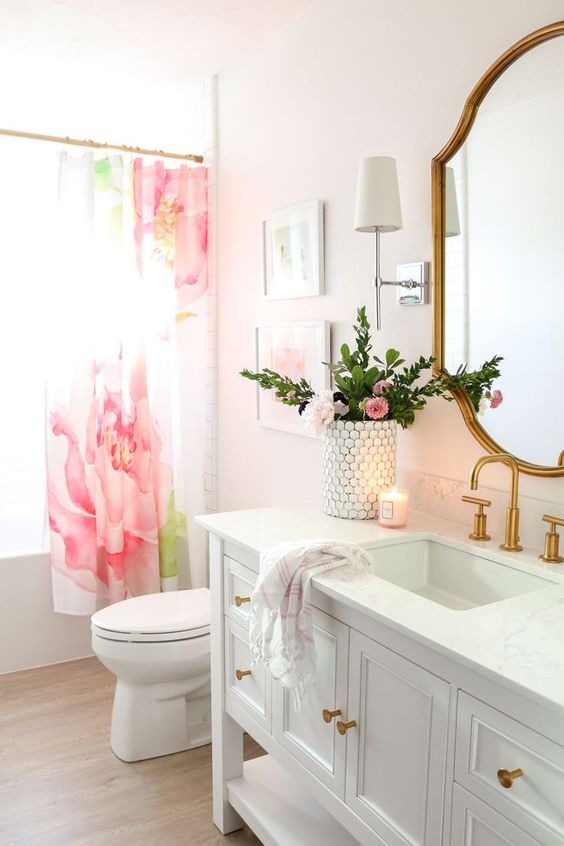 a cute glam bathroom with blush ombre walls, a pink flower curtain, gold fixtures and a chic gold frame mirror plus pink blooms