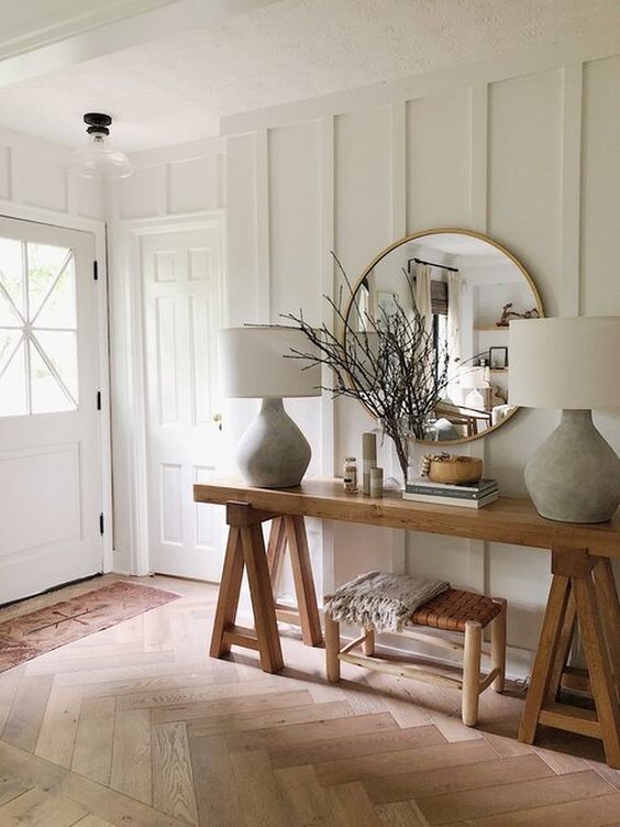 a farmhouse entryway with white paneled walls, wooden furniture, chic lamps and much natural light