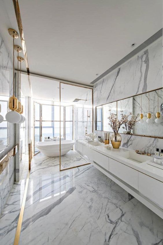 a glam white bathroom all done with marble, with pendant gold lamps, gold frames and a mirror in a gold frame