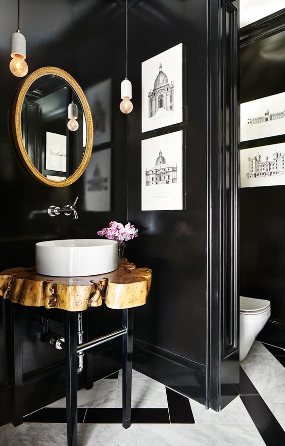 a gorgeous bathroom with a mosaic tile floor, black walls, a gold frame mirror, a wood slice vanity and artworks