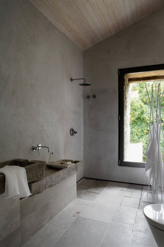 A grey wabi sabi bathroom with concrete walls, grey tiles, stone sinks and a large window to enjoy the views