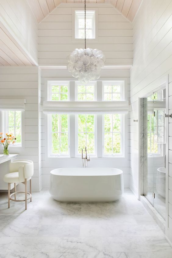 a large white barn bathroom clad with planked walls, a tub by the window, a vanity with a creamy chair and a bubble chandelier