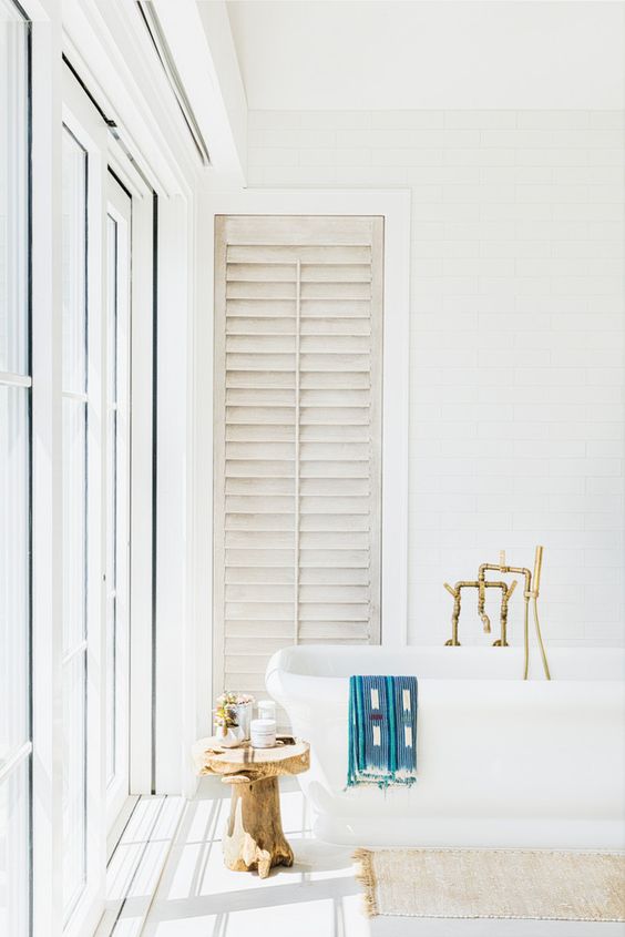 A light filled white bathroom with a free standing tub, brass fixtures, a wooden stool and a glazed wall plus shutters