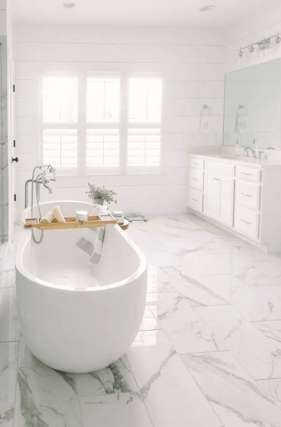 A light filled white bathroom with a large vanity by the window, a white marble tile floor and an oval tub plus potted greenery