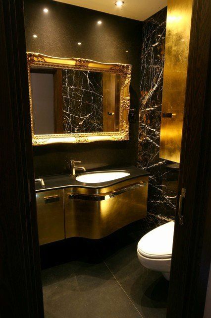 A luxurious powder room with black walls, chic gold touches, a gold vanity, some built in lights and white appliances