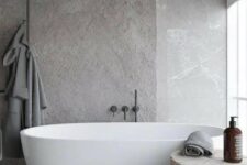 a minimalist and refined grey bathroom fully clad with stone, with a large tree stump side table and an oval tub is chic