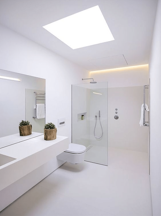 A minimalist white bathroom with a skylight and built in lights, a sleek vanity, a shower with a glass partition and white appliances