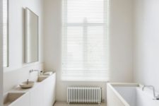 a minimalist white bathroom with a tub and sinks clad with white plywood, mirrors, a radiator and shades on the window