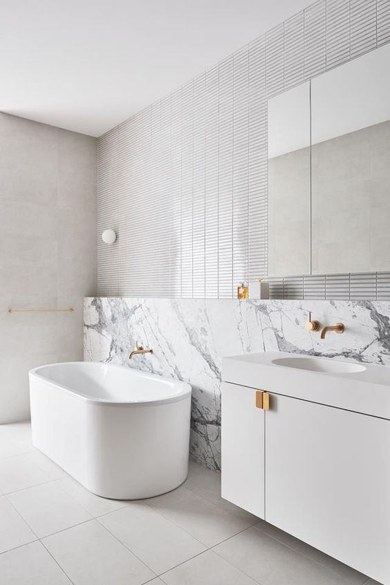 a minimalist white bathroom with stacked tiles and large scale tiles, a sleek vanity, a tub, a mirror on the wall and gold fixtures