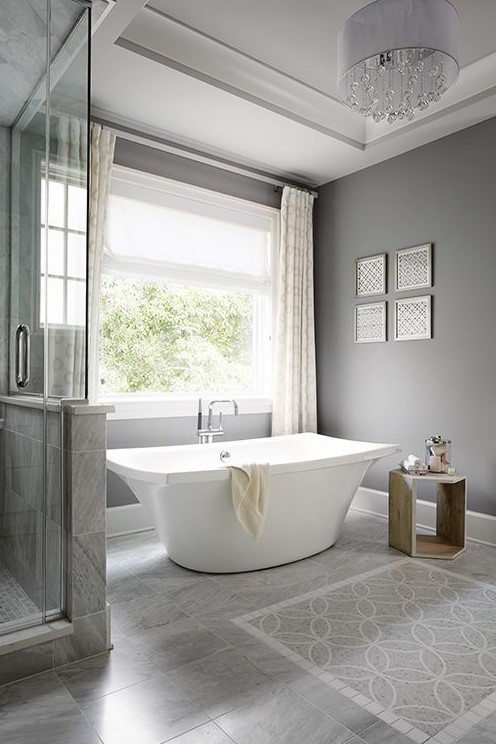 a modern grey bathroom done with tiles on the floor, a catchy free-standing tub, a wooden table and a crystal chandelier