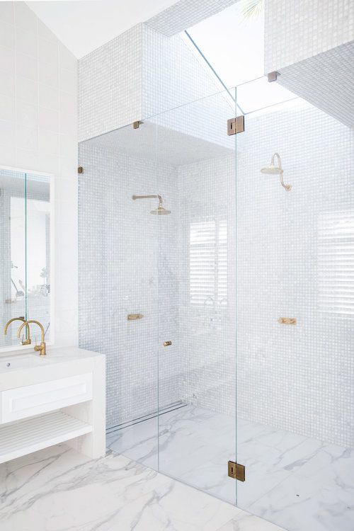 a modern white bathroom with small tiles and marble tiles, a skylight over the shower, a white vanity and gold fixtures