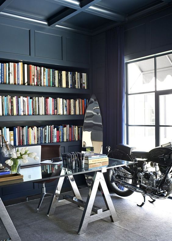 A navy home office with a coffered ceiling, built in storage space, a glass and metal desk plus a bike as decor