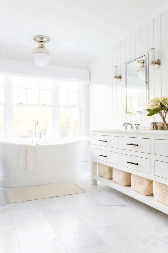 a neutral farmhouse bathroom with a tub by the window, a large vanity, baskets for storage, white marble tiles