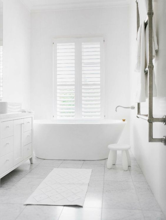 A pure white bathroom with a free standing tub, a small stool, a large vanity and marble tiles on the floor