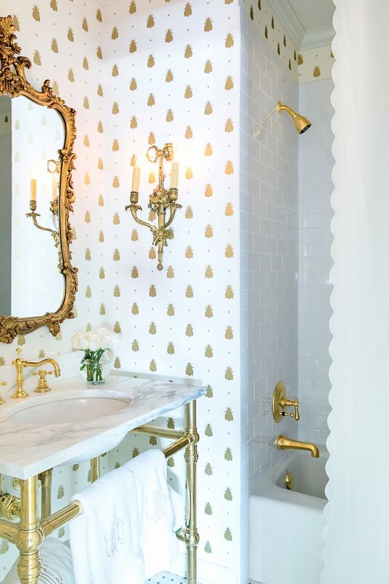 a refined bathroom with white and gold pineapple wallpaper, godl fixtures, a vanity and an exquisite mirror in a gold frame