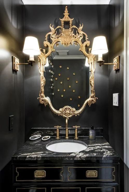a refined black and gold bathroom with a chic ornate mirror, wall sconces, a black vanity and a white sink
