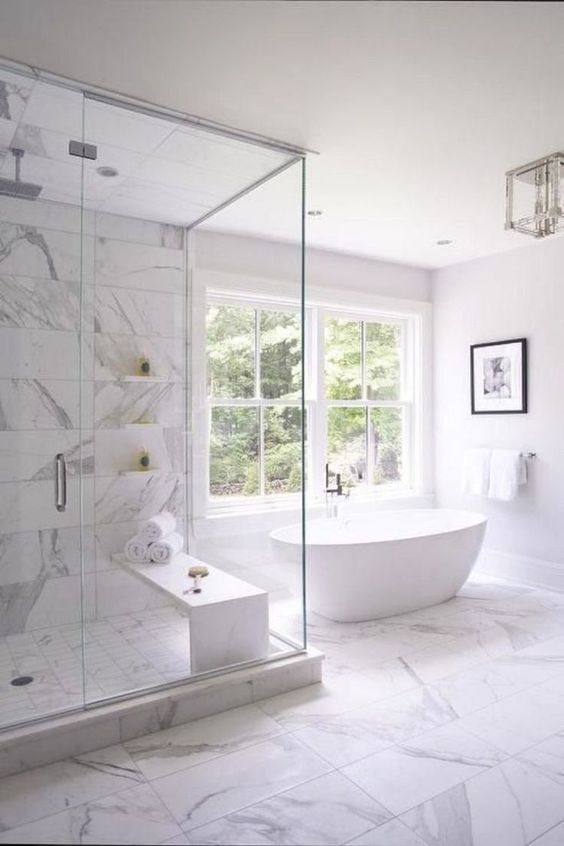a refined white bathroom with marble tiles, a tub by the window, a large shower space, a chic chandelier and some art