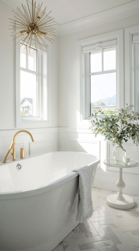a refined white bathroom with windows, a tub, gold fixtures and a gold chandelier and some greenery