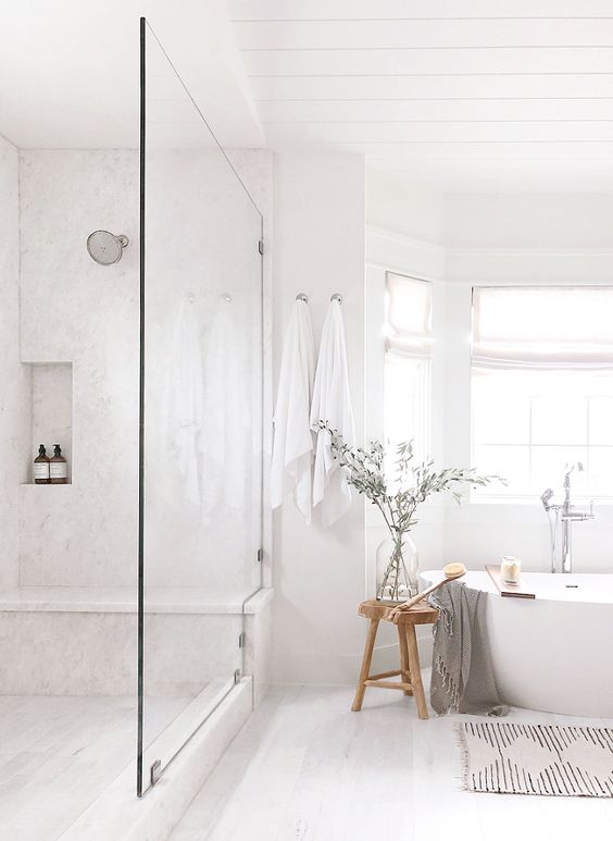 a serene white bathroom with a shower space, a tub by the bay window, a rug, a stool and some greenery