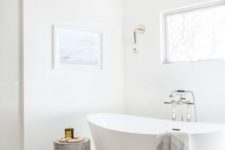 a serene white bathroom with marble tiles on the floor, a tub, a wooden stool, a window and some wall sconces