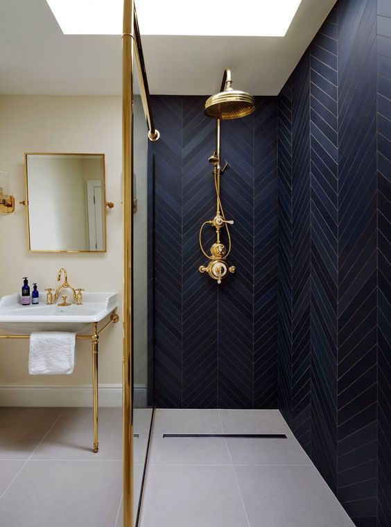 a shower clad with navy tiles in a herringbone pattern, with gold fixtures and gold lamps for a chic look