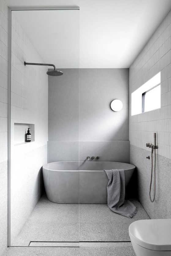 a simple minimalist bathroom clad with skinny white tiles and grey stone-like ones, a concrete tub, white appliances