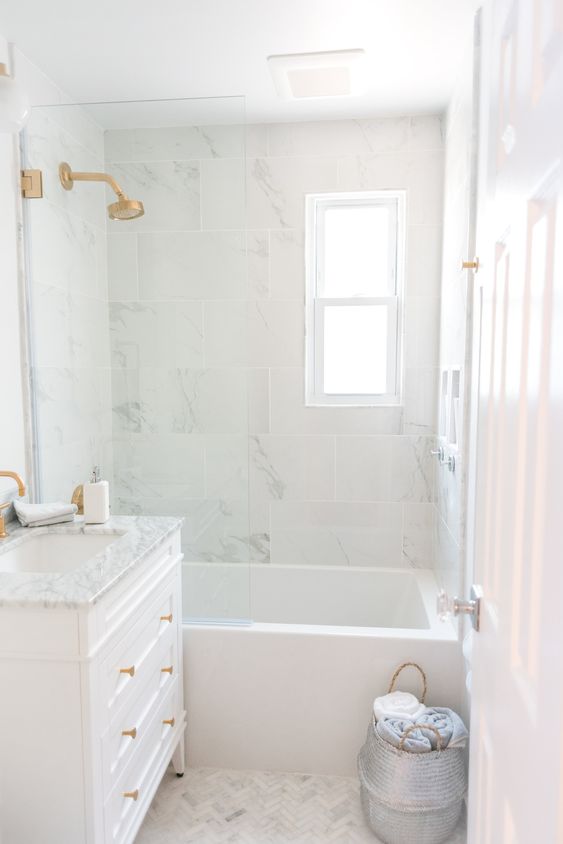 a small and cool white bathroom done with marble tiles, a white vanity, gold fixtures, a basket with towels