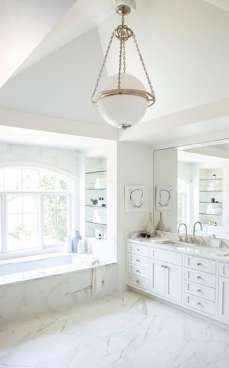 A stylish and elegant bathroom in white, with marble tiles, a large vanity, a tub and built in shelves, a pendant lamp