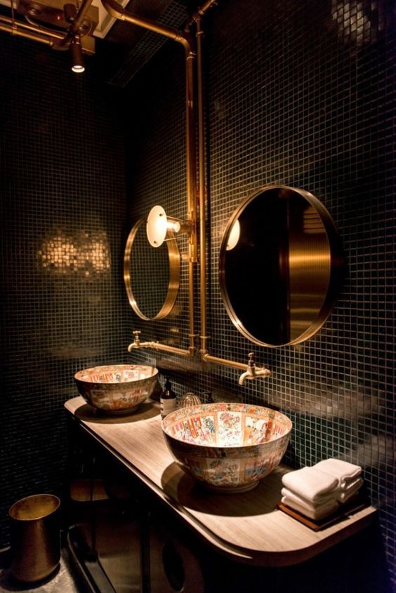 A stylish bathroom with black tiles, gold mirrors, painted sinks and exposed gold pipes is jaw dropping