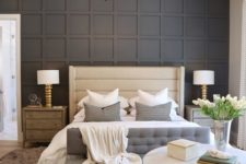 a stylish bedroom with a black paneled wall, an upholstered bed and bench, a round chandelier, wooden nightstands and layered rugs