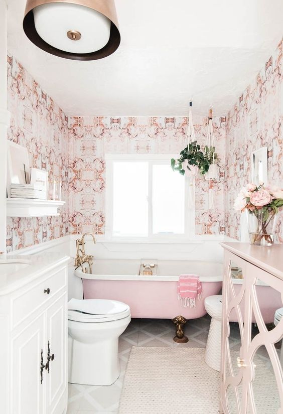 A stylish vintage inspired bathroom done with pink printed wallpaper, a pink bathtub, a pink sideboard and brass fixtures and legs