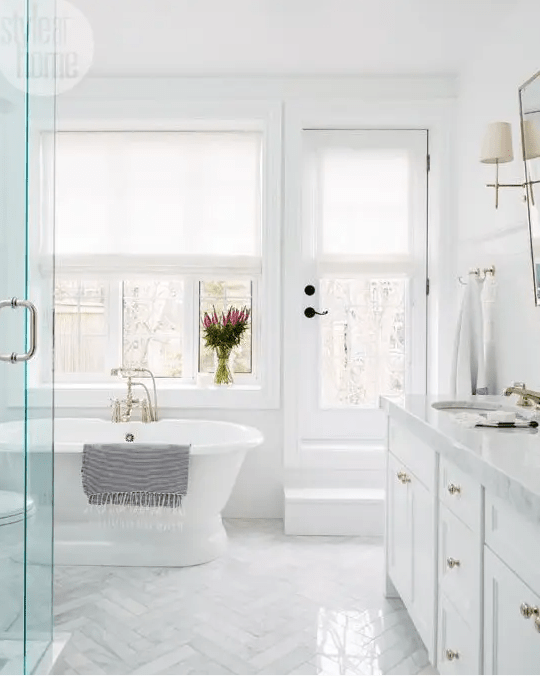 a traditional white bathroom with marble tiles clad in a chevron pattern, a tub by the window and a large vanity