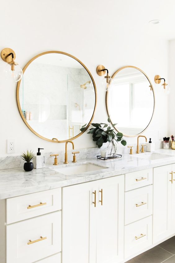 a white bathroom with a large double vanity, a marble countertop, godl framed mirrors, handles and sconces looks nice