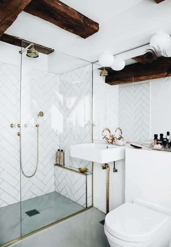a white bathroom with herringbone clad tiles, gold and brass fixtures, dark stained beams and bubble lamps