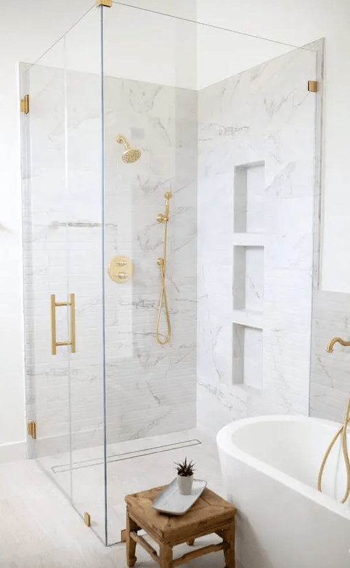 a white bathroom with white marble tiles, a shower space with niches, an oval tub, a wooden stool and gold fixtures