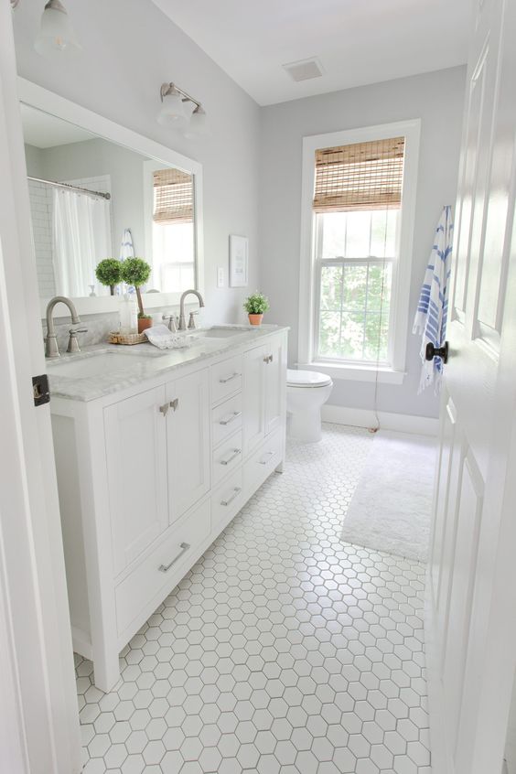 a white farmhouse bathroom with hex tiles, a vanity, a shower space, woven shades and some greenery