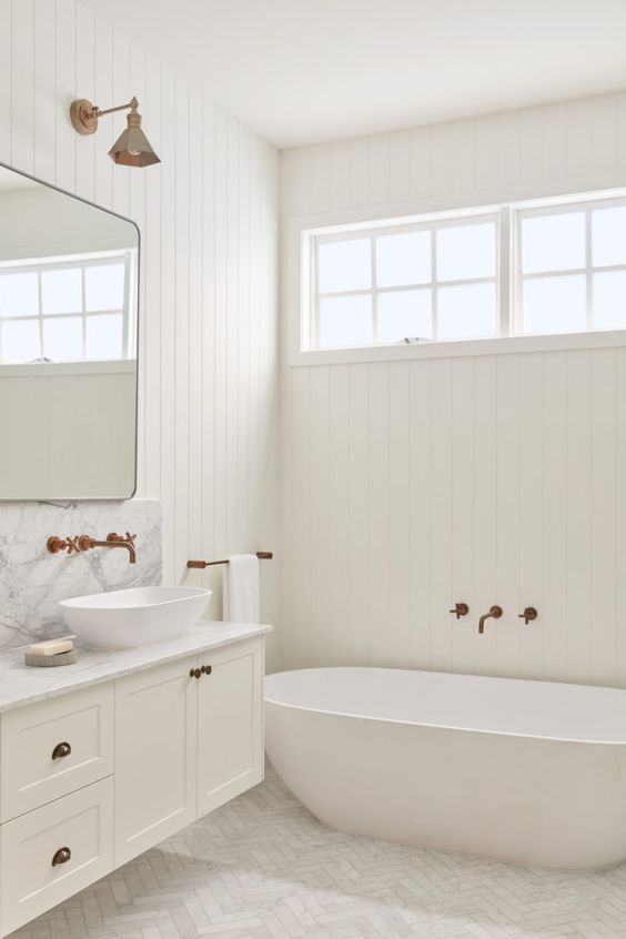 a white farmhouse bathroom with planked walls, marble tiles, an oval tub, a floating vanity and brass and copper fixtures