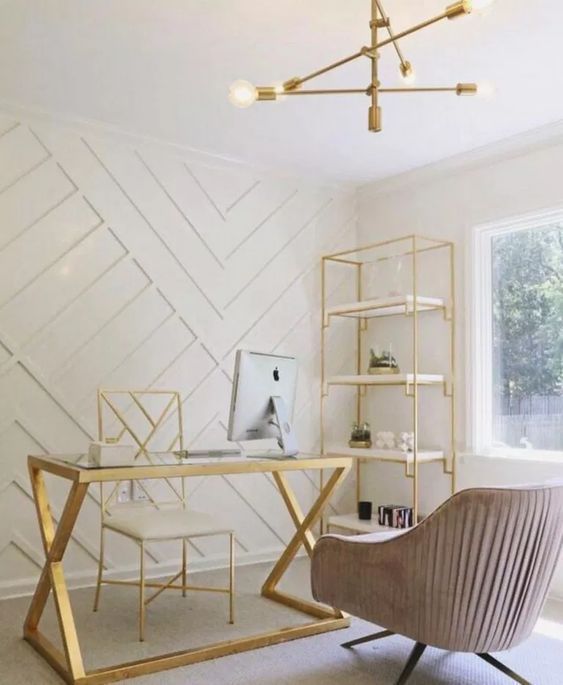 an airy feminine home office with a white paneled wall, gold furniture, a stylish chandelier and a pink chair