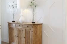 an elegant farmhouse entryway with a white paneled wall, a wooden sideboard and chandelier, a white lamp and greenery