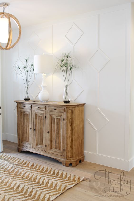 an elegant farmhouse entryway with a white paneled wall, a wooden sideboard and chandelier, a white lamp and greenery