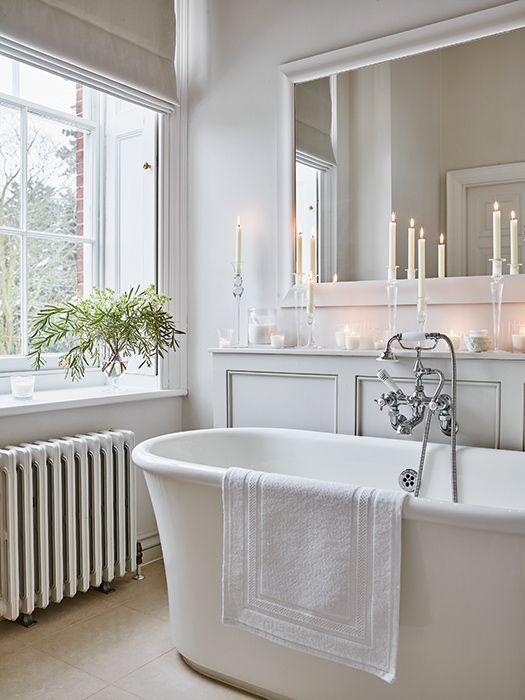 an elegant white bathroom with a paneled console, a tub, candles, a large mirror and a radiator under the window