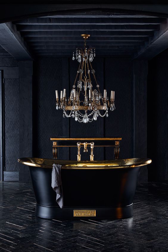 an exquisite black and gold bathroom clad with tiles and done with textural wallpaper, with a black tub and a chic chandelier with crystals