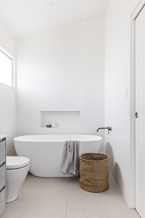 an ultra-minimalist white bathroom with a window, a tub, a niche, a vanity, a basket and dark fixtures is cool and clean