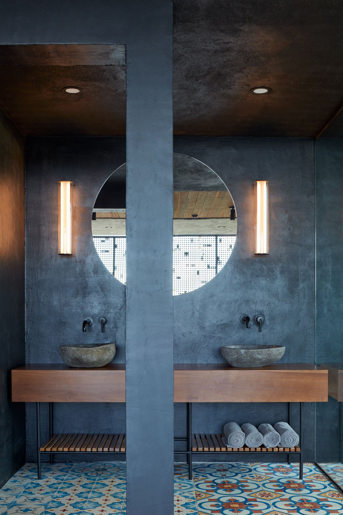 The master bathroom shows off concrete walls, cool  colorful tile floors, and rough stone sinks