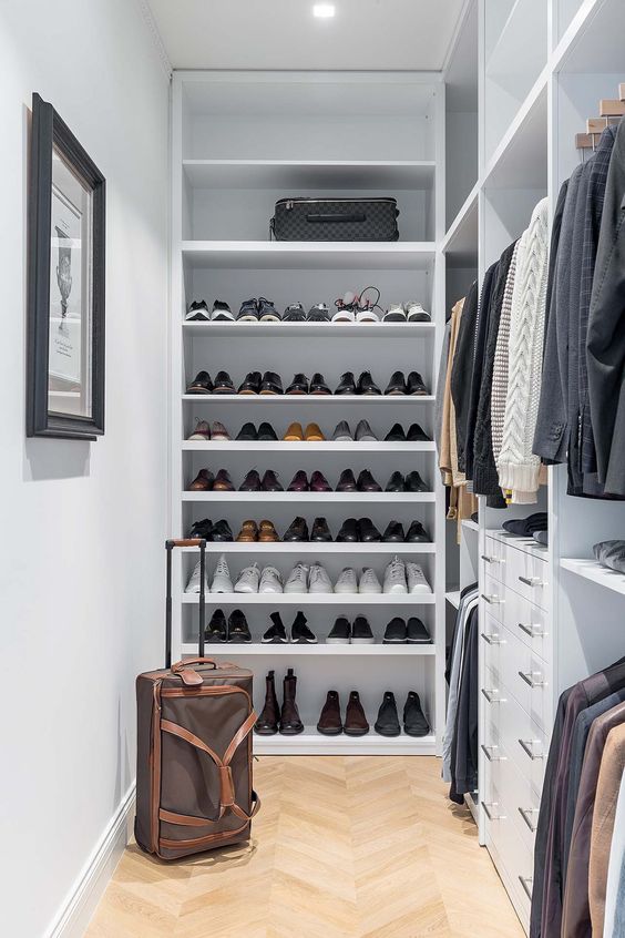 A small and stylish manly closet in white, with lots of open shoe shelves, holders with hangers, built in drawers and lights