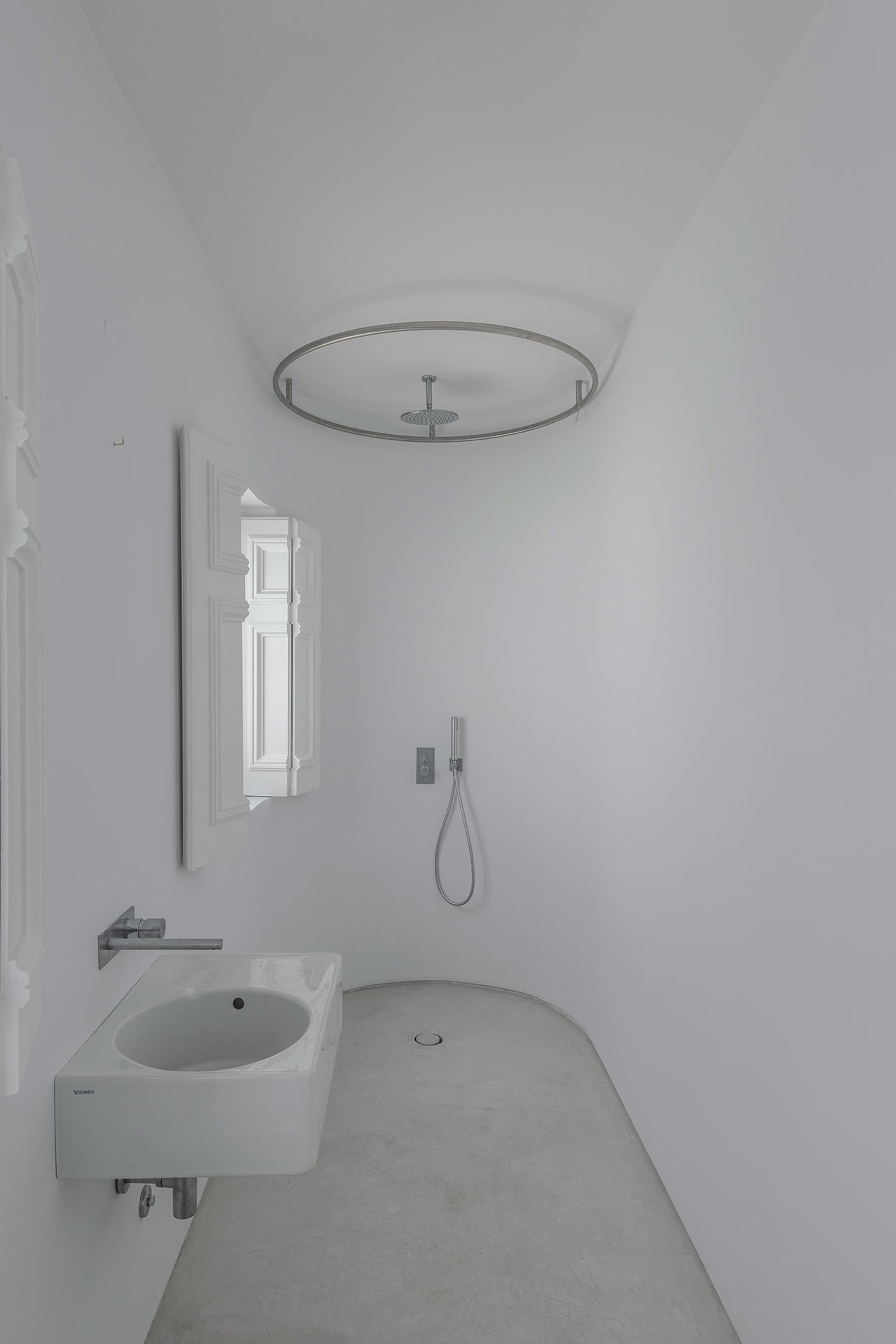 The bathroom is pure white, with a small shower space and a wall mounted sink, the original shutters preserved